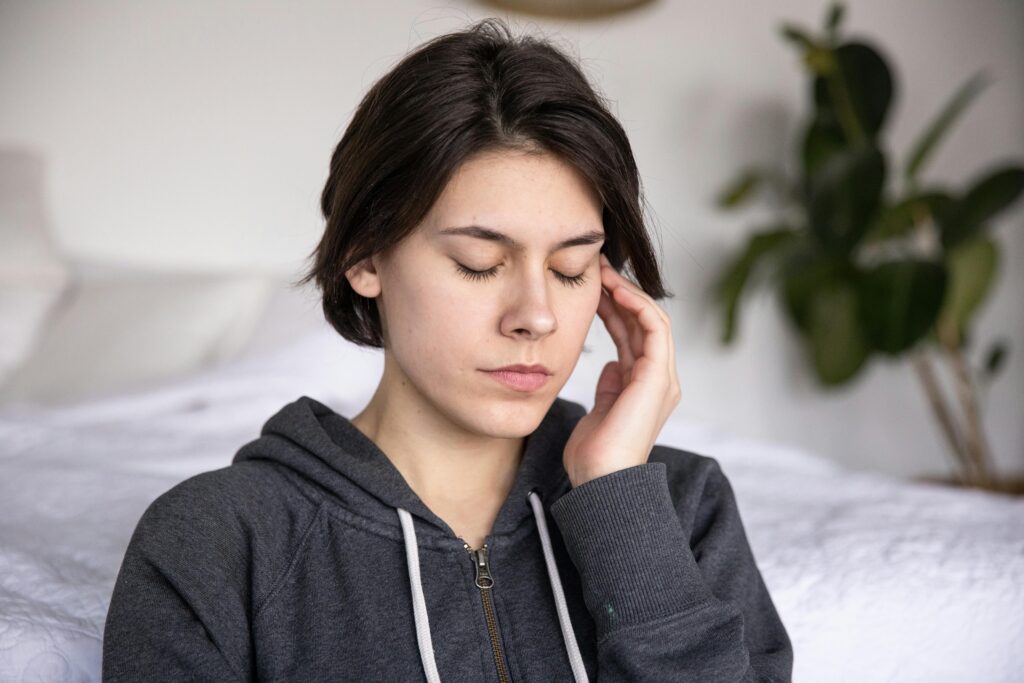 A young woman with closed eyes sits thoughtfully indoors, wearing a hoodie, suggesting deep reflection or meditation.
